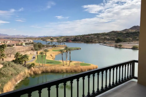 Scenic view from a balcony overlooking a lake, golf course, and mountains under a partly cloudy sky.