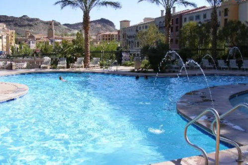 A clear blue swimming pool surrounded by palm trees and buildings, with a person swimming in the distance.
