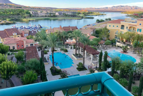 Aerial view of a resort with pools, palm trees, and a river, surrounded by mountains and colorful buildings.