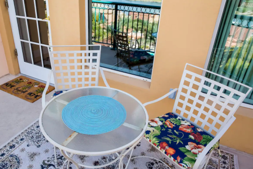 A small outdoor seating area with a round table and two chairs, featuring floral cushions and a reflection in the window.