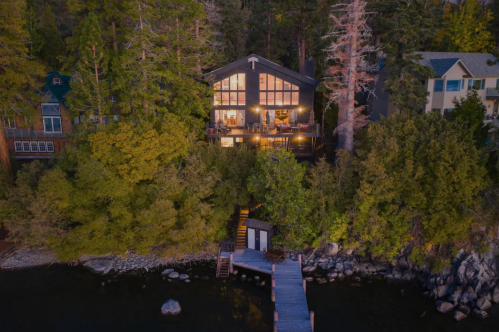 A modern cabin by a lake, surrounded by trees, with large windows reflecting the evening light. A dock extends into the water.