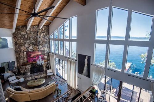 Spacious living room with large windows overlooking a serene lake, featuring a stone wall and modern decor.