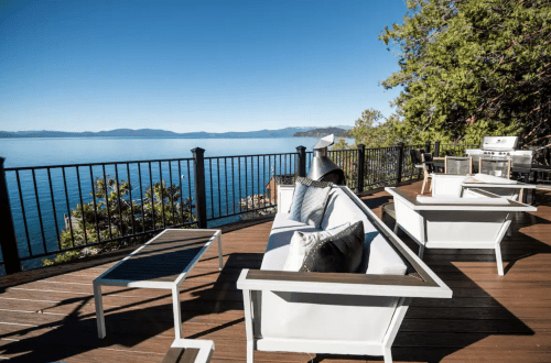 A scenic deck with modern furniture overlooking a calm lake and distant mountains under a clear blue sky.