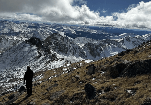 A hiker stands on a rocky, snow-covered mountain slope, overlooking a vast landscape of peaks and valleys under a cloudy sky.