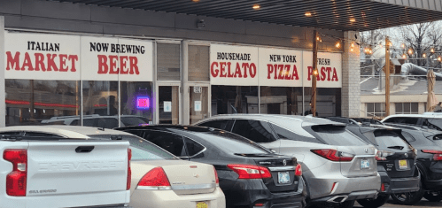 A storefront with signs for Italian market, beer, gelato, pizza, and fresh pasta, with parked cars in front.