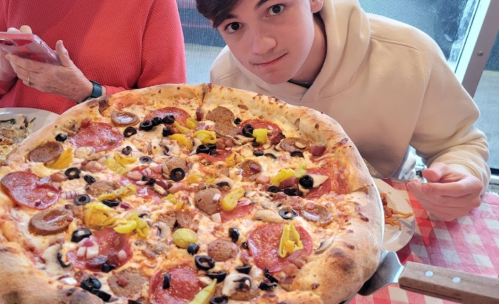 A young person in a hoodie poses with a large pizza topped with pepperoni, olives, and various other toppings.
