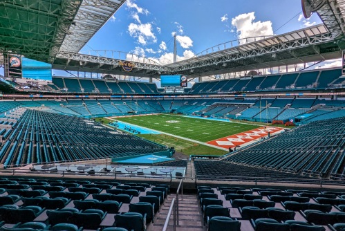 A wide view of a modern stadium with empty blue seats and a green field under a bright sky.
