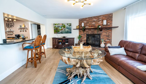Cozy living room with a glass table, leather sofa, and a brick wall, featuring a TV and bar seating area.