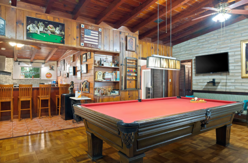 A cozy game room featuring a red pool table, wooden walls, a bar area, and various wall decorations.