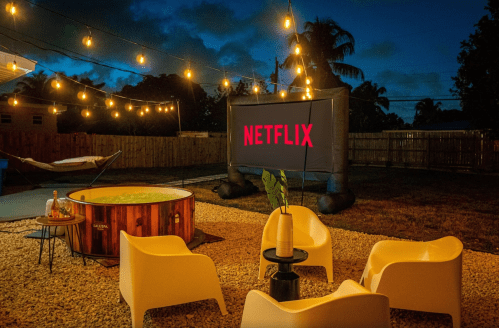 Outdoor movie setup with a Netflix screen, hot tub, and cozy seating under string lights at dusk.