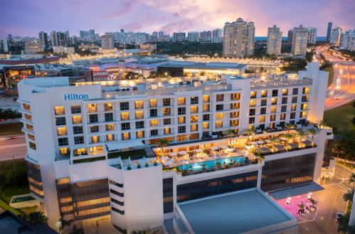 Aerial view of a Hilton hotel with a rooftop pool, surrounded by city skyline and evening lights.