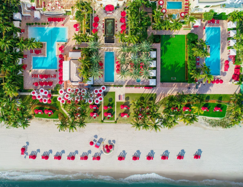 Aerial view of a resort with pools, palm trees, and beach chairs along a sandy shore, featuring red umbrellas and loungers.