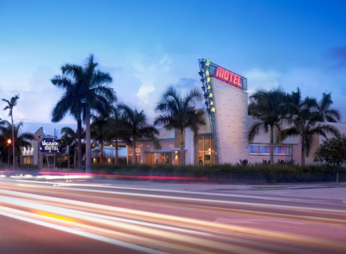 A retro-style motel with neon signage, surrounded by palm trees, at dusk with light trails from passing cars.