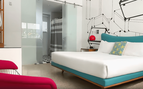 A modern hotel room featuring a large bed, colorful decor, and a bathroom with a glass door in the background.