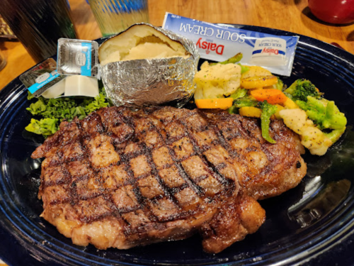 A grilled steak with a baked potato, sour cream, and mixed vegetables on a blue plate.