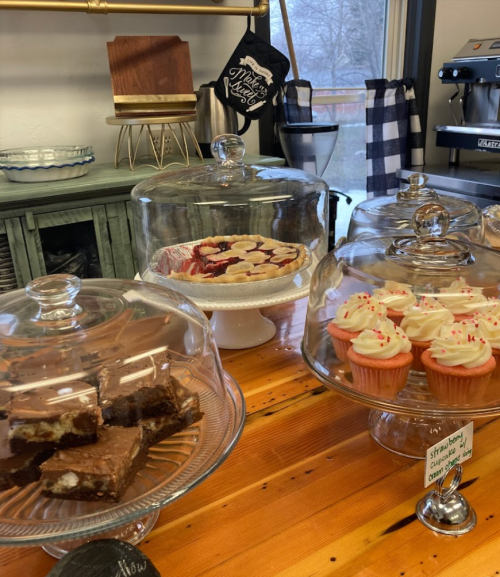 A display of desserts under glass domes, including brownies, a fruit tart, and cupcakes with sprinkles on a wooden table.