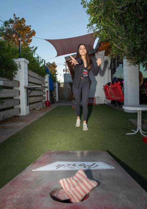 A woman in athletic wear throws a bean bag towards a cornhole board in a lively outdoor setting.