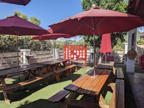 Outdoor seating area with wooden picnic tables, red umbrellas, and a Connect Four game in a sunny garden setting.