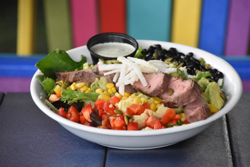 A colorful salad bowl with grilled meat, corn, black beans, tomatoes, avocado, and a side of dressing.