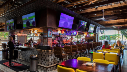 A vibrant restaurant interior featuring a bar with multiple TVs, colorful decor, and patrons seated at tables.