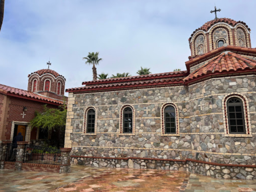 A stone church with domed roofs and crosses, surrounded by palm trees and a cloudy sky.