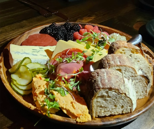A wooden platter with assorted cheeses, meats, pickles, bread, and fresh berries.