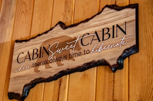 Wooden sign reading "Cabin Sweet Cabin" with a bear silhouette, featuring the phrase "where there's always time to hibernate."