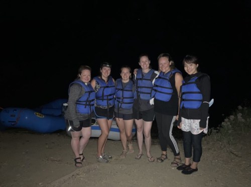 A group of six women in life jackets stand together at night near a blue raft.
