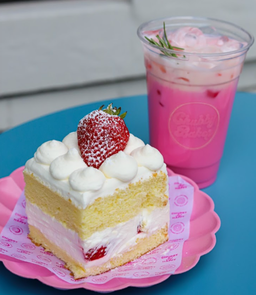 A slice of strawberry cake with whipped cream and a drink in a clear cup, featuring pink layers and a strawberry garnish.