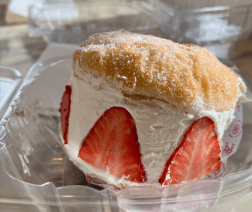 A fluffy dessert with whipped cream and fresh strawberries, topped with powdered sugar, in a clear plastic container.