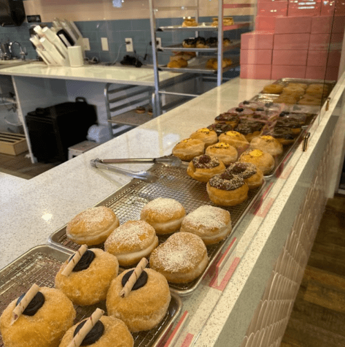A display of assorted donuts on trays, featuring various toppings and fillings, in a bright bakery setting.