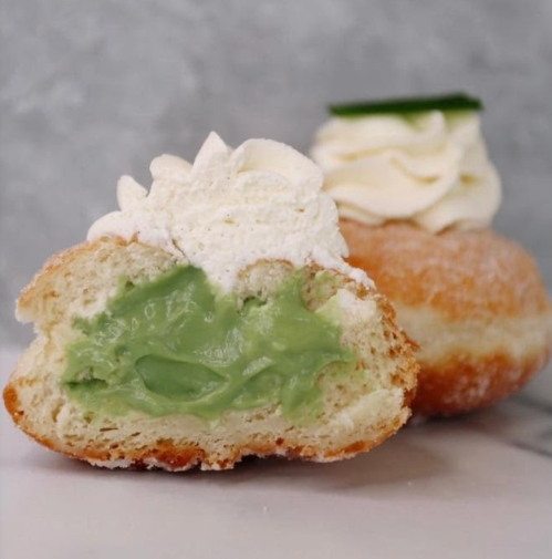 A split donut filled with green cream and topped with whipped cream, alongside another donut with a green topping.