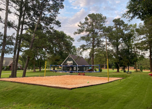A sandy volleyball court with a blue house and tall trees in the background, set in a green yard.