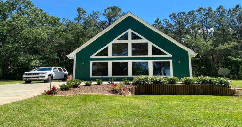 A green house with large windows and a triangular roof, surrounded by a lawn and flower beds, with a white SUV parked nearby.