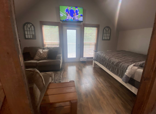 Cozy room with a bed, sofa, wooden table, and a TV, featuring large windows and wooden flooring.