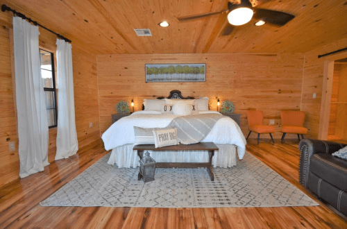 Cozy bedroom with wooden walls, a large bed, decorative pillows, and two armchairs, featuring warm lighting and a rug.