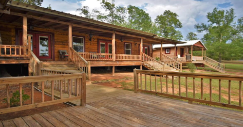 A rustic wooden cabin with a porch, surrounded by trees and a grassy area, under a partly cloudy sky.