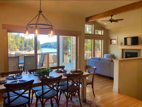 Bright dining area with a wooden table, chairs, and a view of a lake through large windows. Cozy living space in the background.