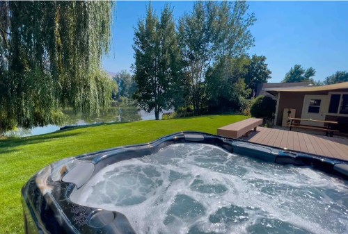 A hot tub with bubbling water overlooks a serene lake and lush green lawn under a clear blue sky.