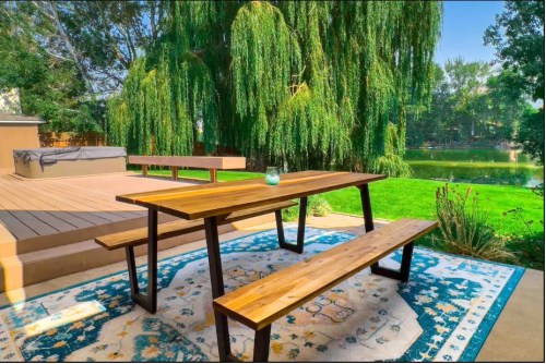 A wooden picnic table and benches on a patio, surrounded by greenery and overlooking a serene pond.