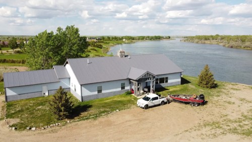 Aerial view of a riverside house with a boat and truck parked outside, surrounded by trees and a scenic landscape.