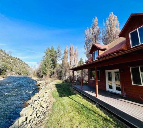 A red cabin by a river, surrounded by trees and mountains under a clear blue sky.