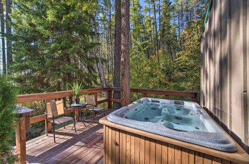 A wooden deck with a hot tub surrounded by tall trees and greenery, featuring two chairs and a small table.