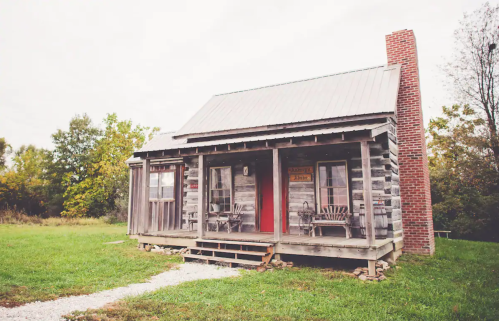 A rustic log cabin with a red door, surrounded by greenery and featuring a porch with wooden chairs.