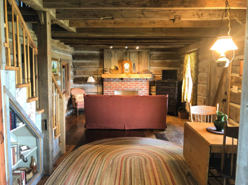 Cozy rustic living room with wooden beams, a brick fireplace, and vintage furniture, featuring a braided rug and warm lighting.
