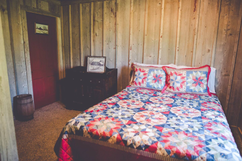 Cozy bedroom with a wooden wall, featuring a quilted bedspread and a red door. A small dresser and barrel are visible.
