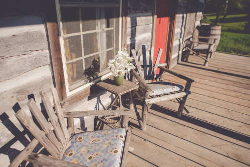 A rustic porch with two wooden chairs, a small table, and a vase of flowers beside a log cabin.