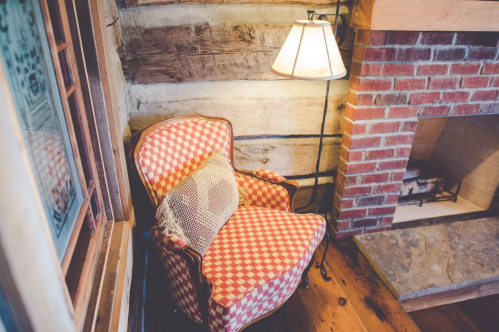 Cozy red and white checkered armchair beside a lamp, against a rustic wooden wall and brick fireplace.