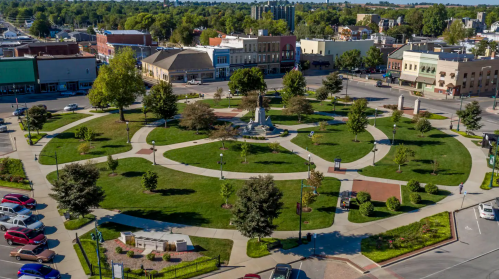 Aerial view of a park with pathways, greenery, and a central monument, surrounded by buildings and cars.