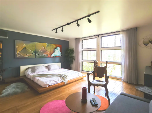 A modern bedroom with a wooden bed, large windows, artwork on the wall, and a cozy seating area.